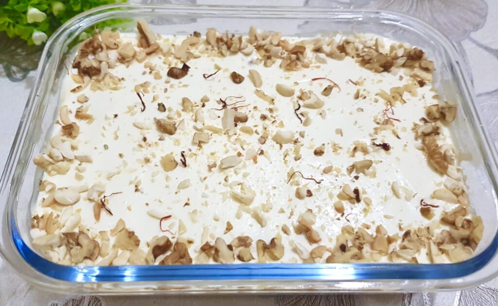 A serving of creamy Arabian pudding garnished with crushed pistachios and a drizzle of rose syrup, presented in a glass bowl on a decorative Middle Eastern-style tray.